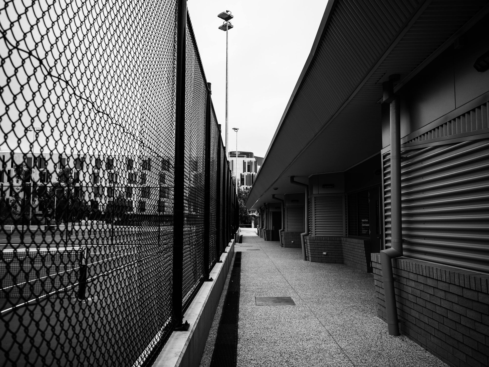 Chain-link fence beside a covered walkway along a brick building, with gravel ground and trees in the distance.