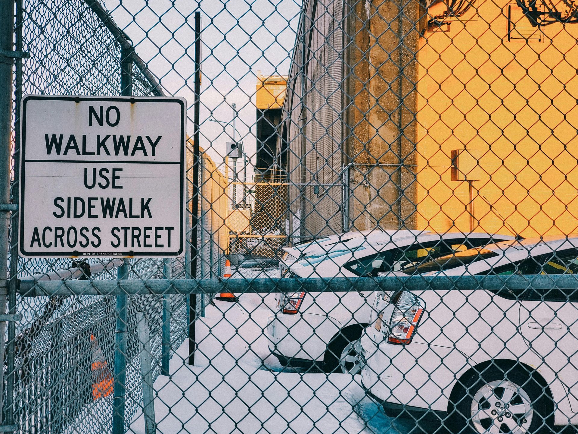 “No Walkway Use Sidewalk Across Street” sign on chain-link fence beside parked cars and snow-covered ground
