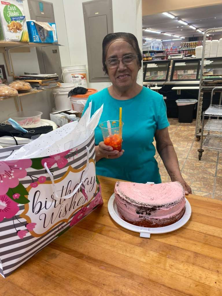 A person wearing a teal shirt stands behind a wooden counter with a pink frosted cake and a birthday gift bag.
