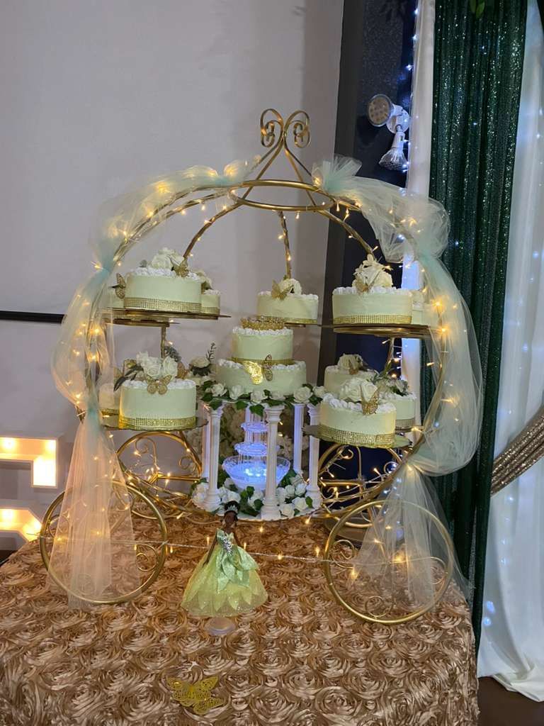 A golden carriage-shaped dessert stand holding multiple small white cakes, illuminated by fairy lights on a gold tablecloth.