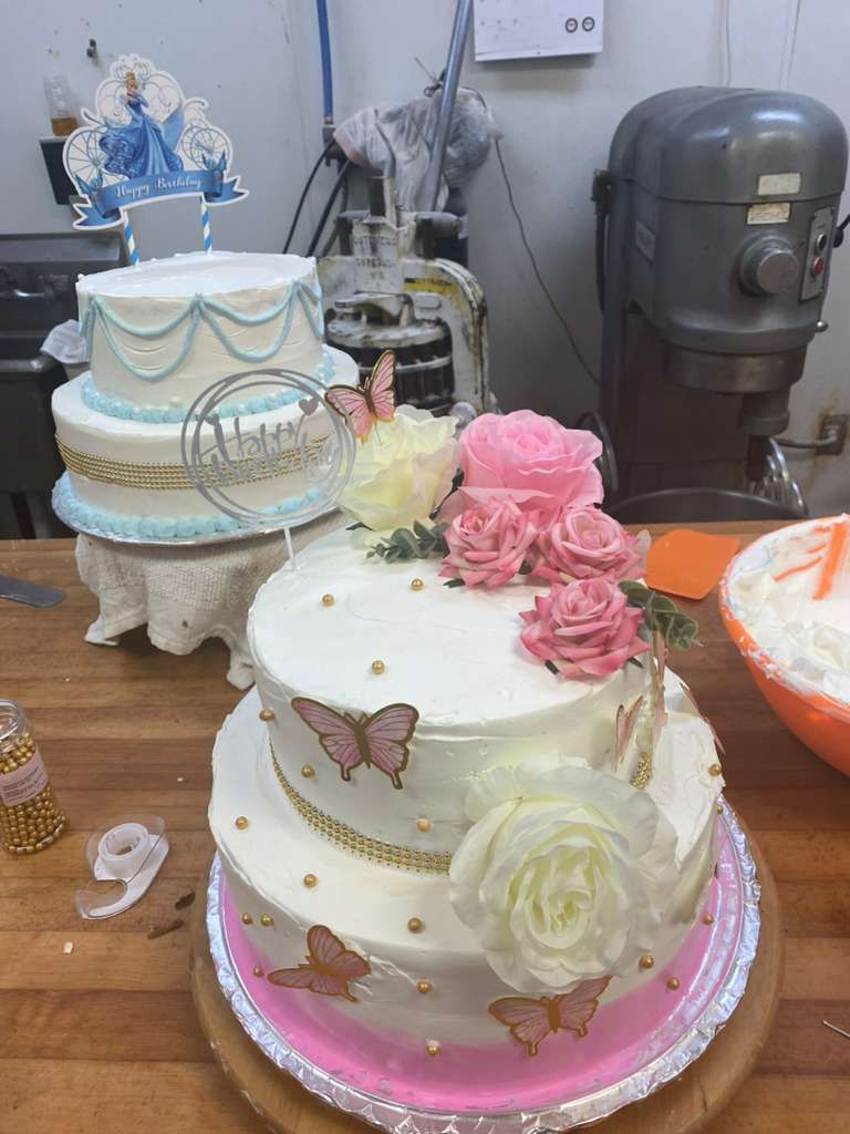 Two tiered cakes decorated with flowers, butterflies, and a Cinderella topper on a bakery workstation.