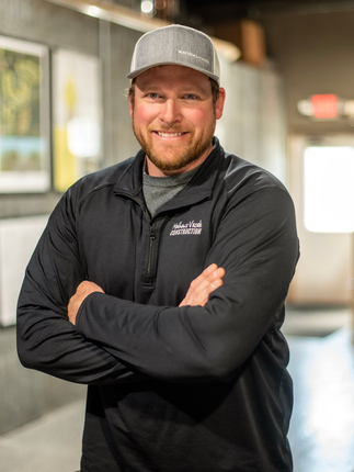Man in black jacket and cap, arms crossed, smiling in hallway.
