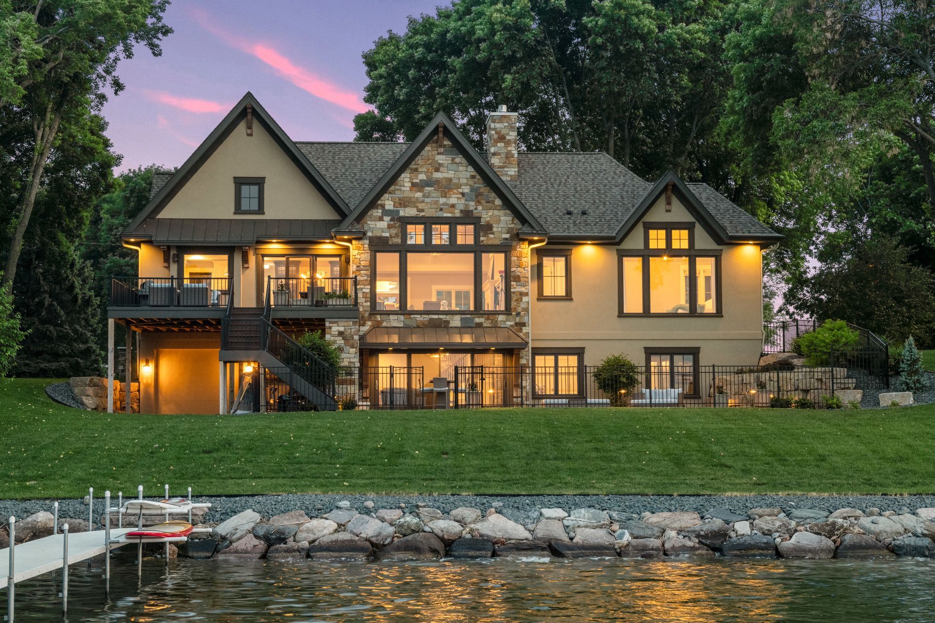 Lakeside house with stone facade, large windows, and manicured lawn at dusk, with dock.
