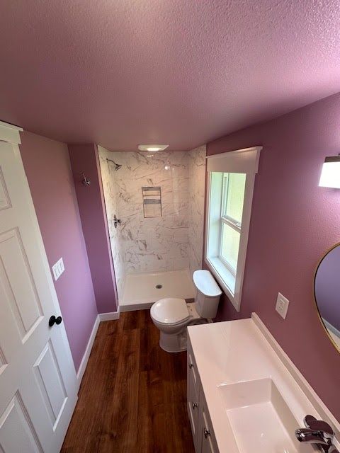 Bathroom with mauve walls, marble shower, white toilet and vanity, and wood-look flooring.