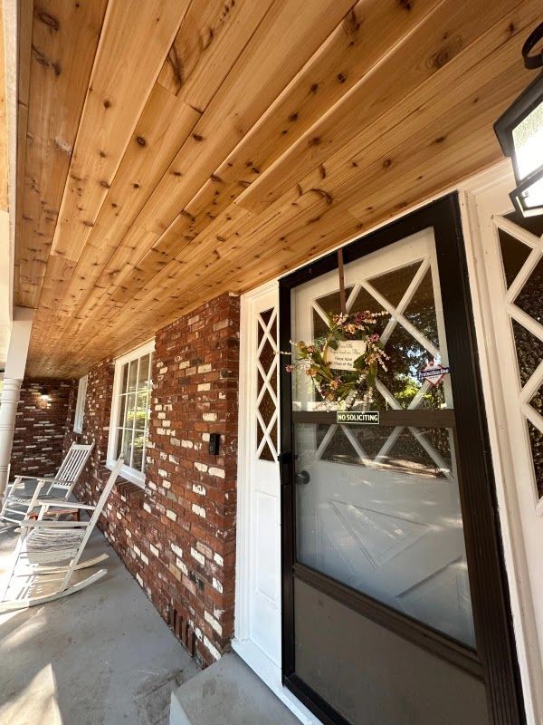 Brick home porch with a wood ceiling and front door. A wreath hangs on the door; rocking chairs are visible.