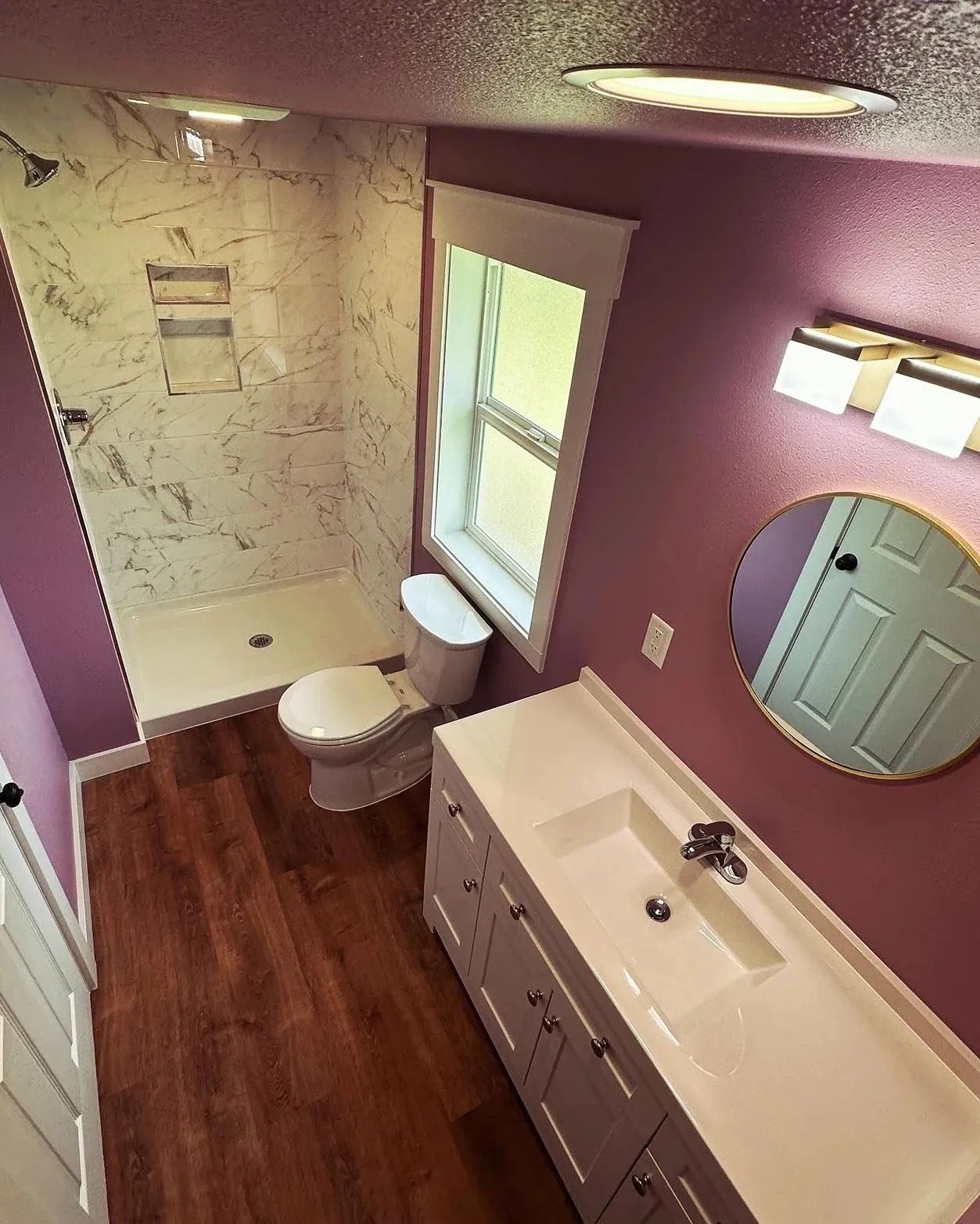 Purple bathroom with white vanity, round mirror, window, toilet, and tiled shower