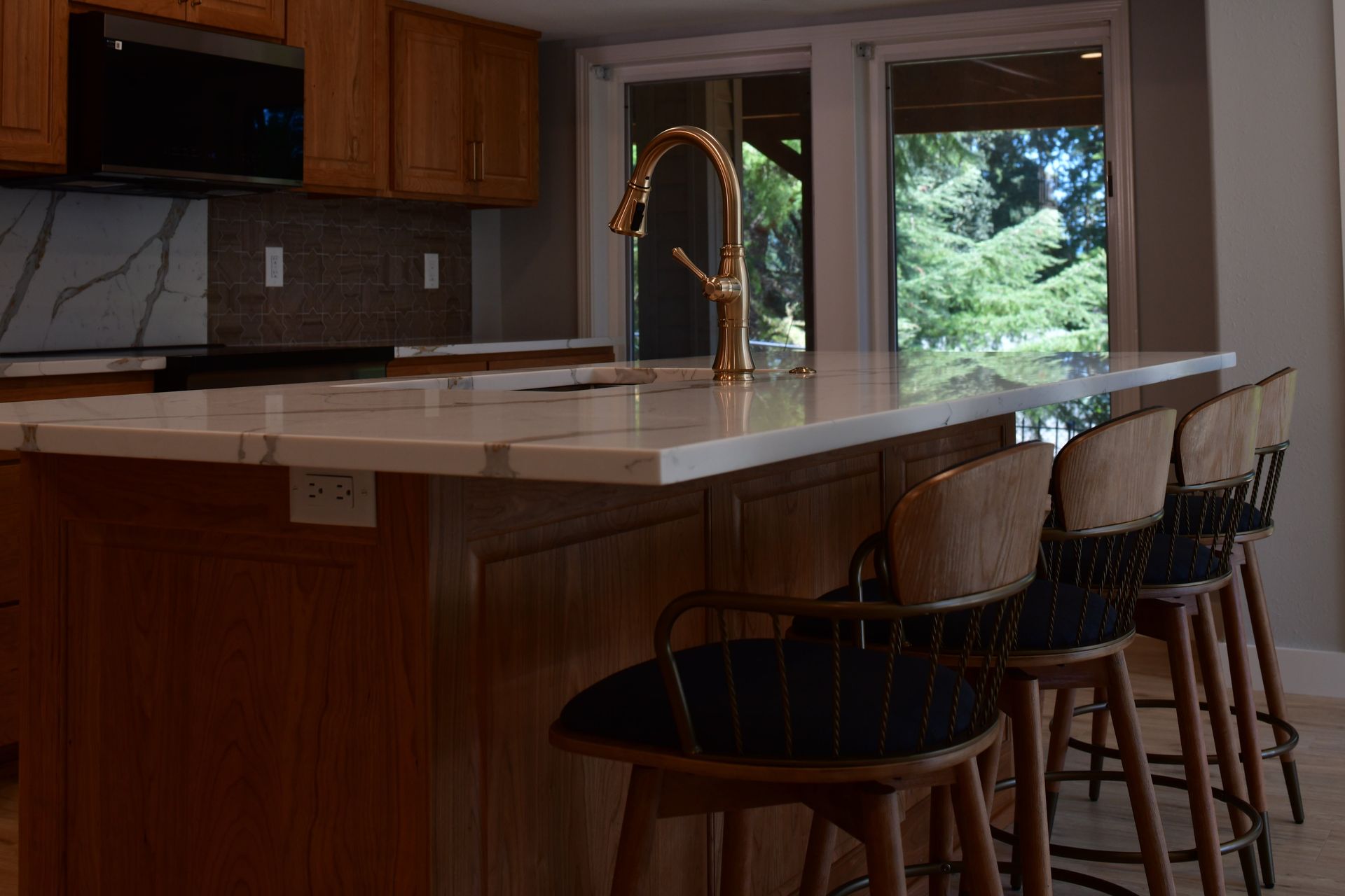 Kitchen with a white countertop island, gold faucet, wooden cabinets, and stools, with a view of trees.