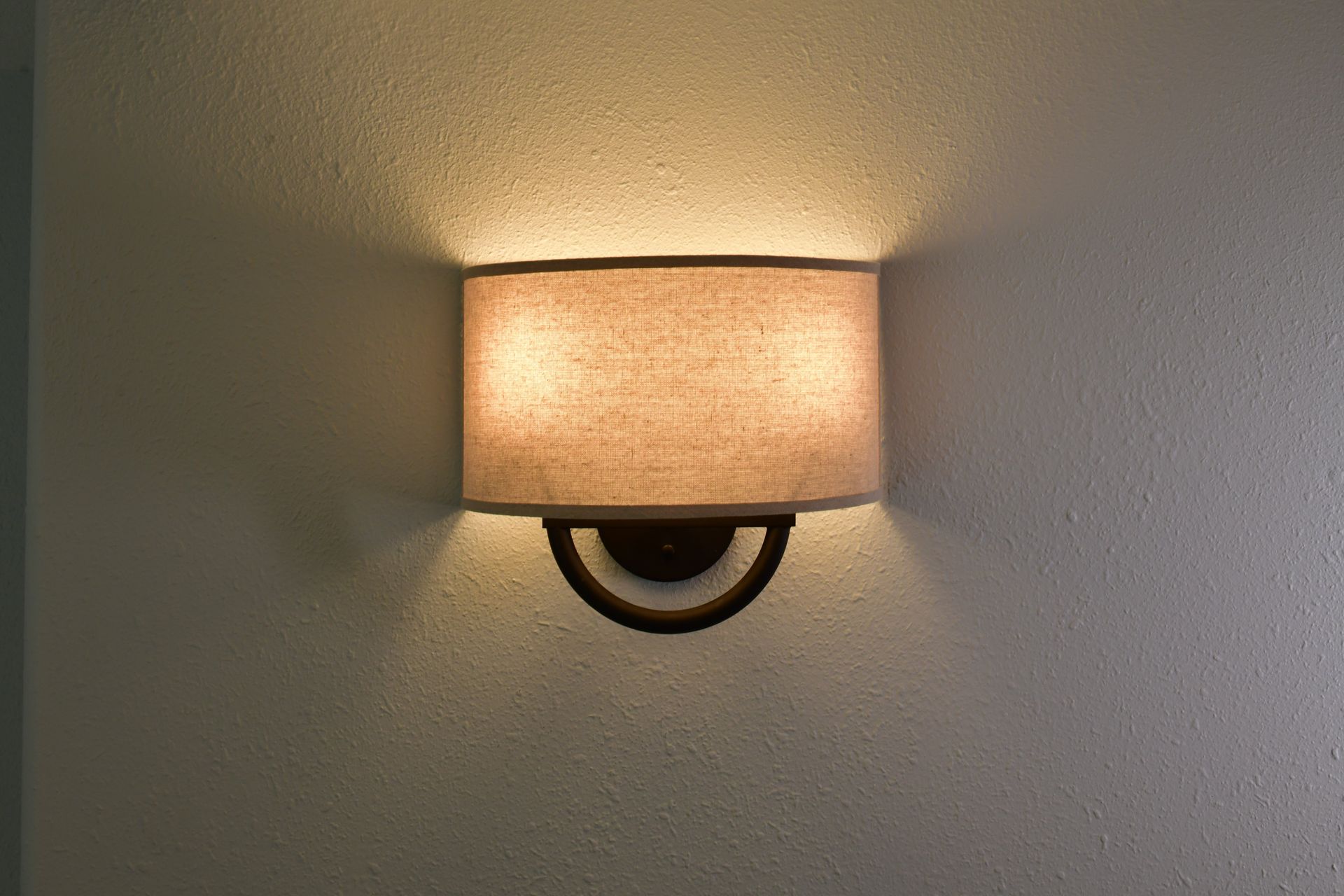 Wall-mounted lamp with a tan lampshade, casting soft light on a textured wall.