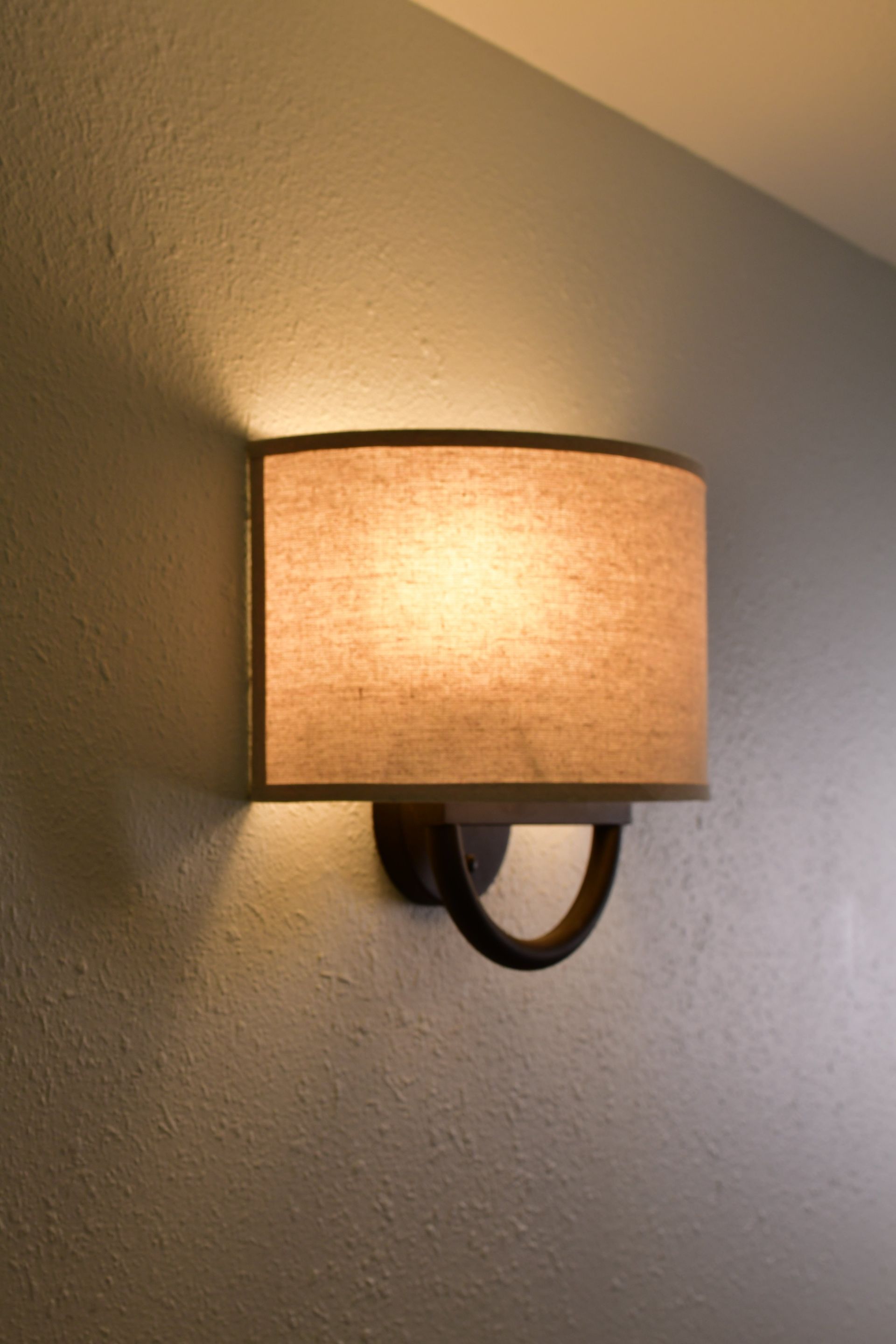 Wall-mounted light fixture with a tan lampshade, casting warm light on textured gray wall.