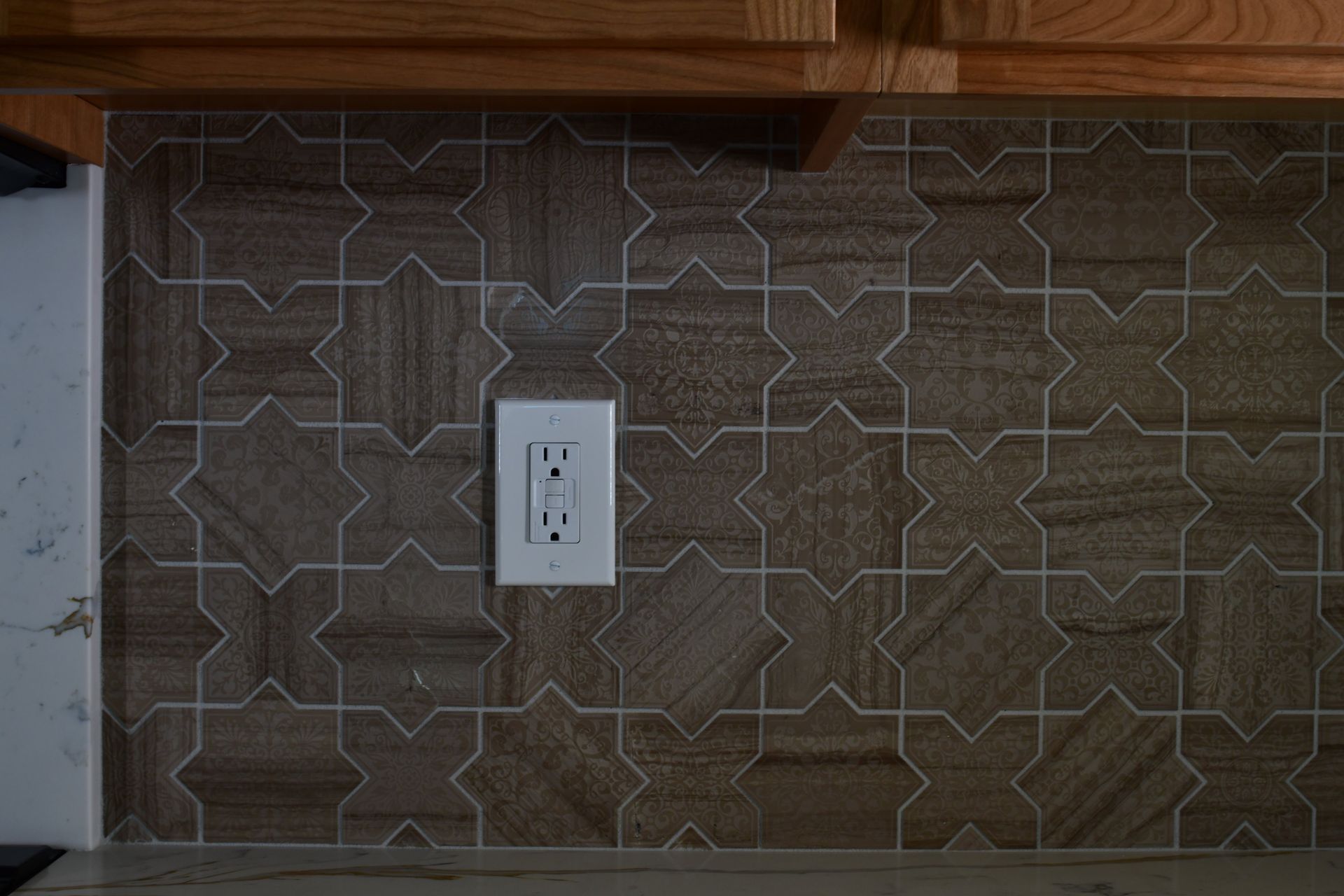Brown star-patterned tile backsplash with a white electrical outlet below wood cabinets.