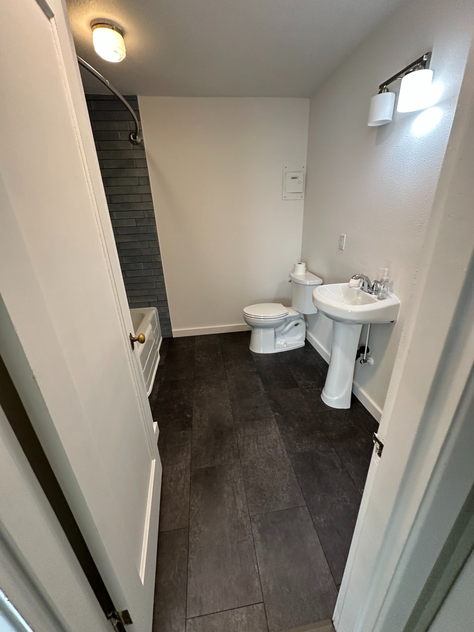 Bathroom with dark wood-look flooring, white fixtures, and shower with dark tile accent wall.