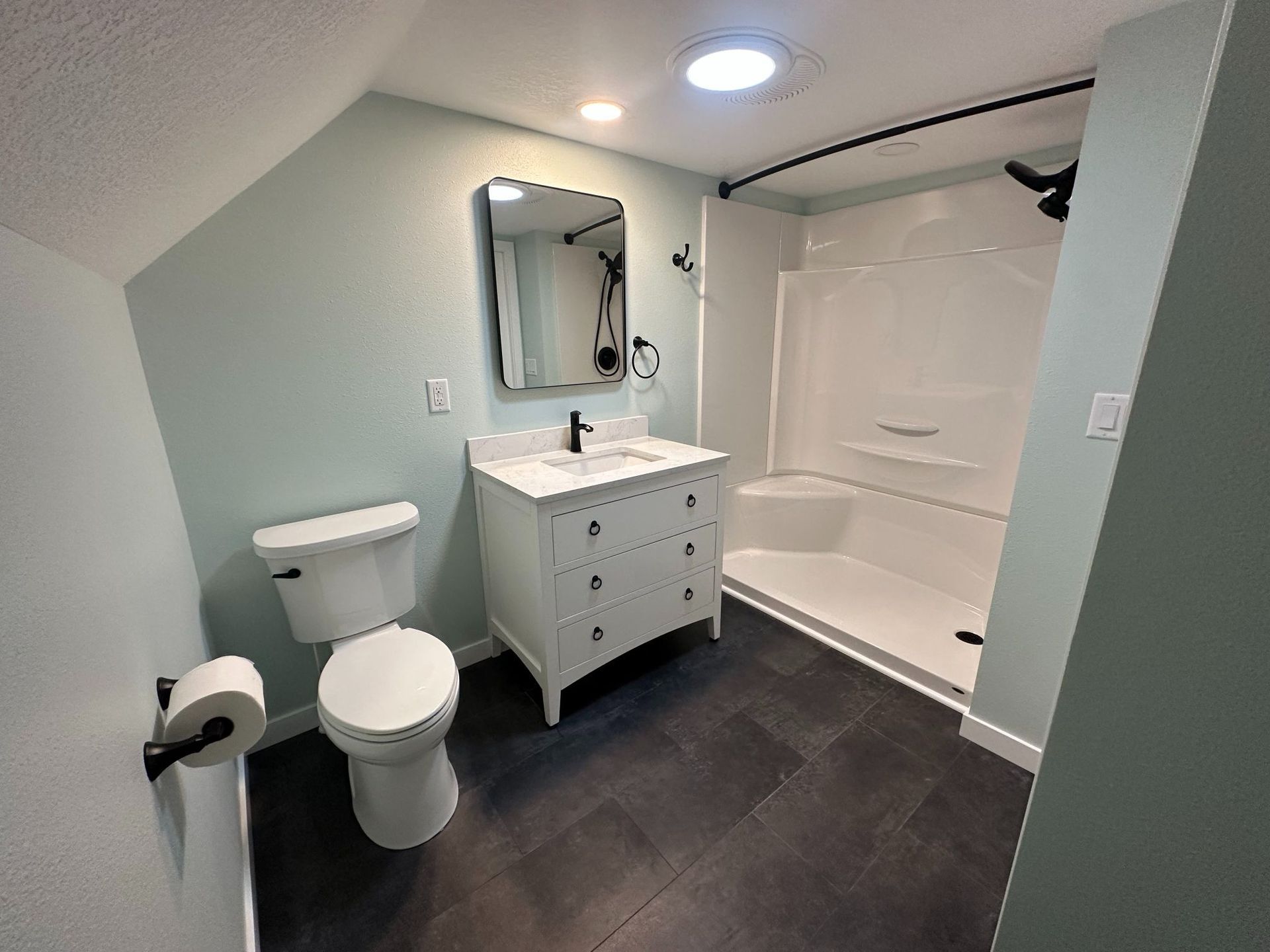 Bathroom with a white vanity, toilet, and shower against a light blue wall; dark floor.