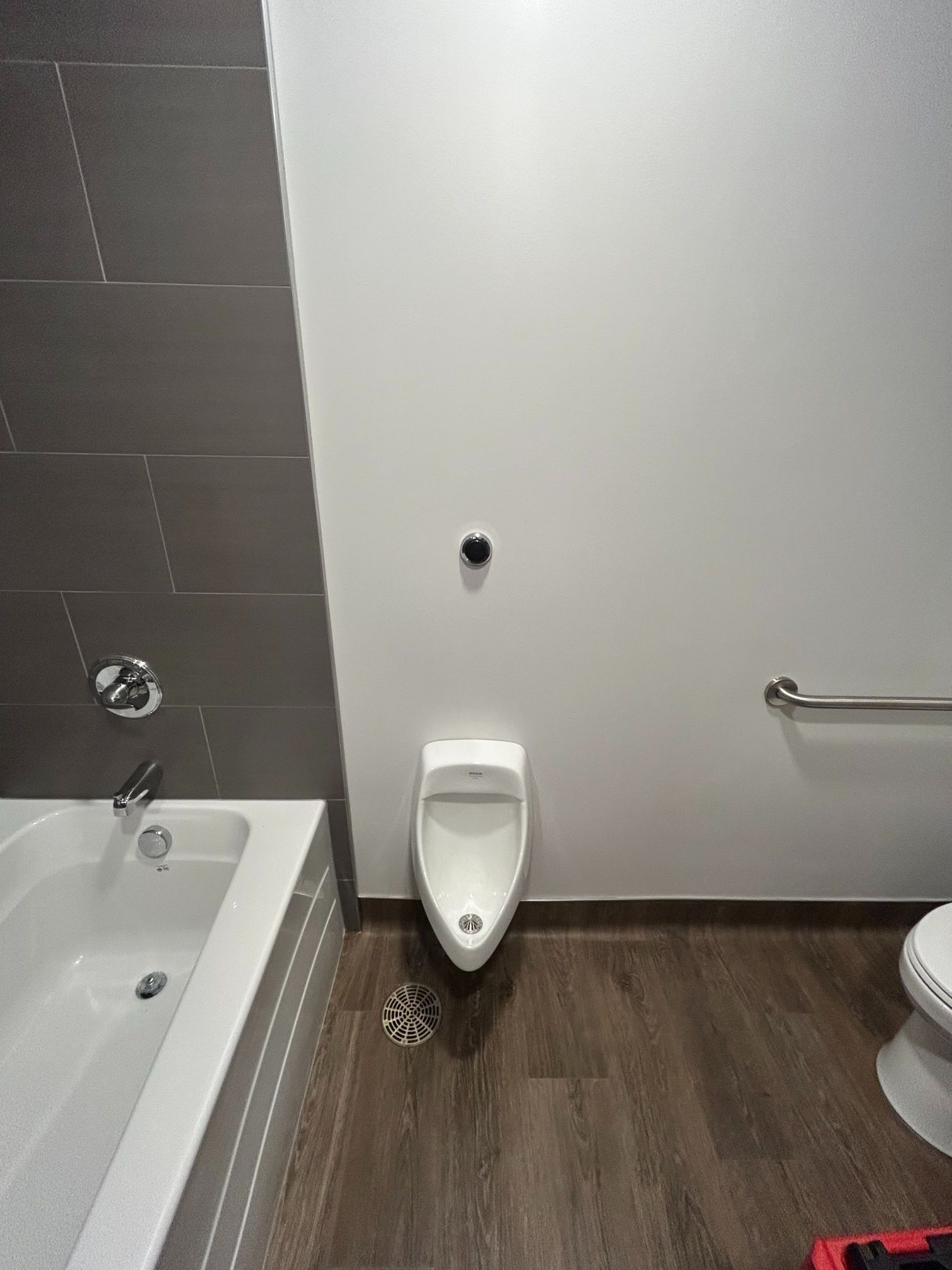 Bathroom with a white urinal, bathtub, and toilet; wood-look floor, gray tile wall, and grab bar.