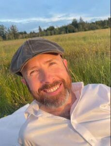 Man with a beard and cap smiles outdoors in a field.