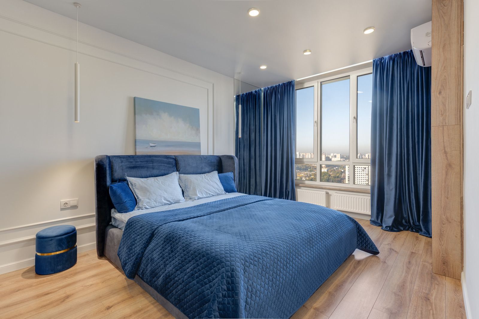 Bedroom with blue velvet bedding, curtains, and stool. Window with city view.