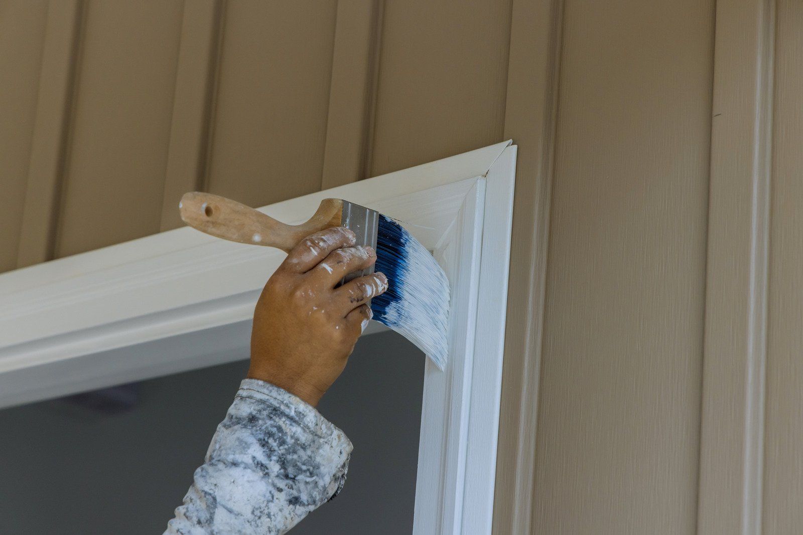Person painting white trim around a door with a paintbrush; beige siding.