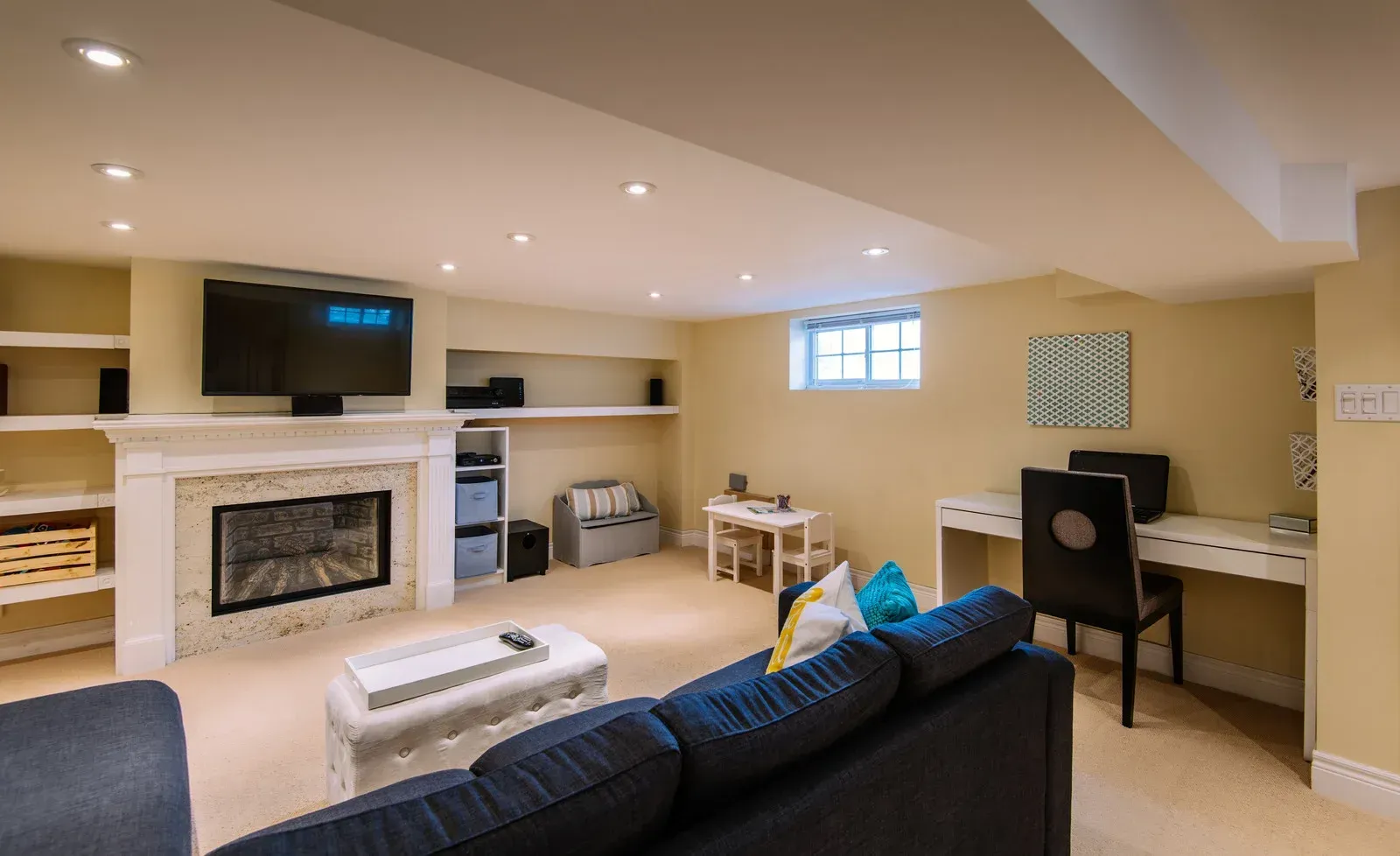 Basement living room with fireplace, sofa, desk, and built-in shelving. Beige walls, cream fireplace, and recessed lighting.