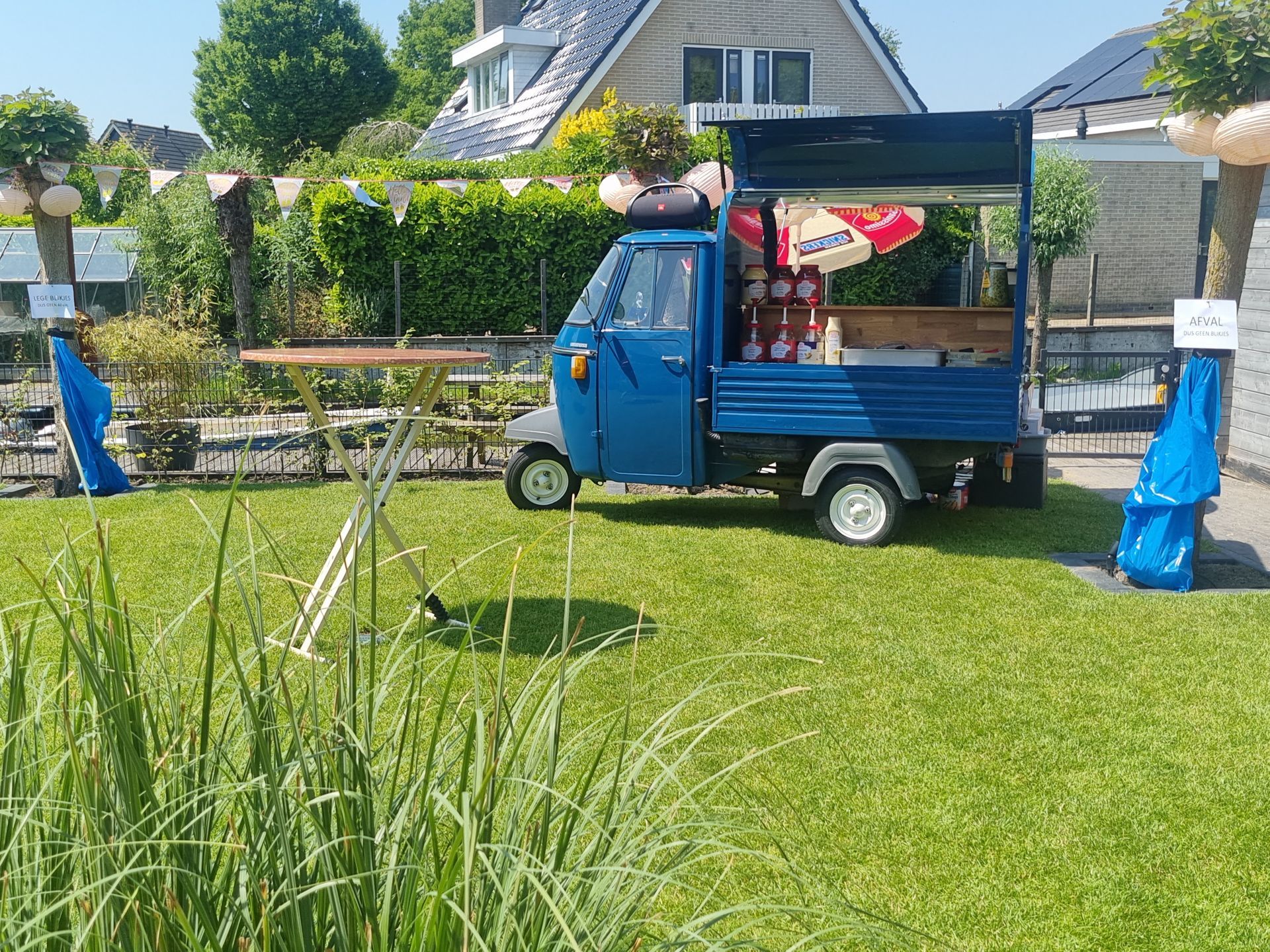 Een blauwe foodtruck staat op een grasveld met mensen die onder een afdak eten serveren, vlakbij huizen en bomen.