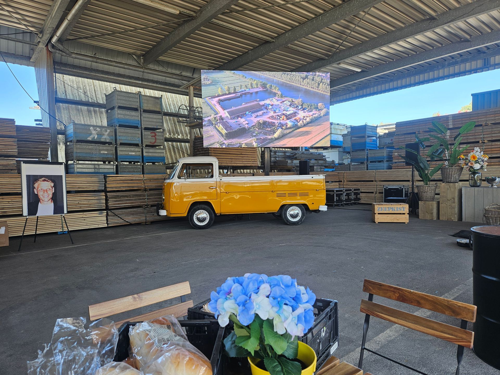 Een overdekte ruimte met een gele volkswagen, een groot led scherm, opgestapelde goederen en blauwe bloemen op de voorgrond.