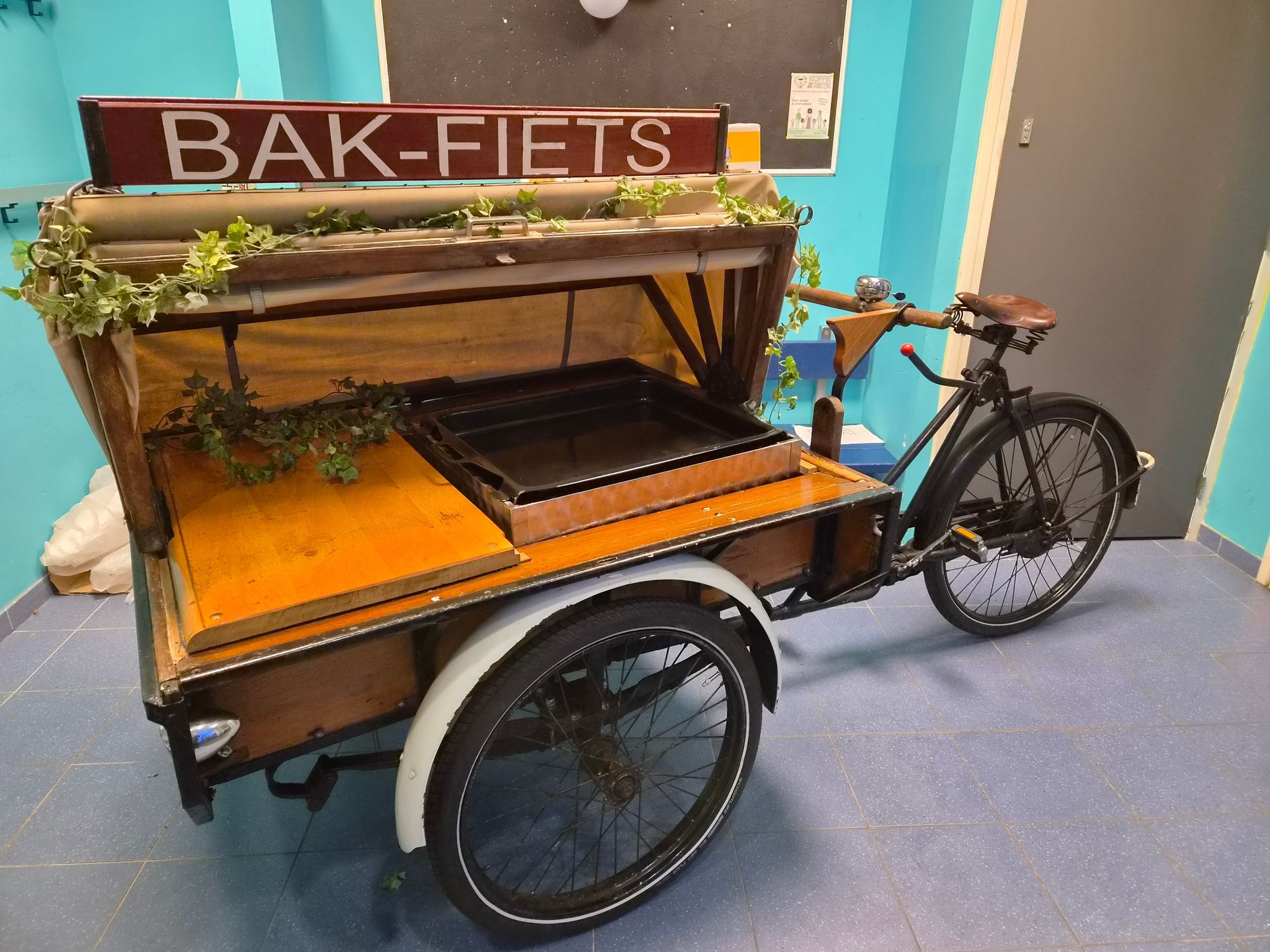 Bakfiets met houten laadbak en luifel, geparkeerd binnen op een blauwe vloer.