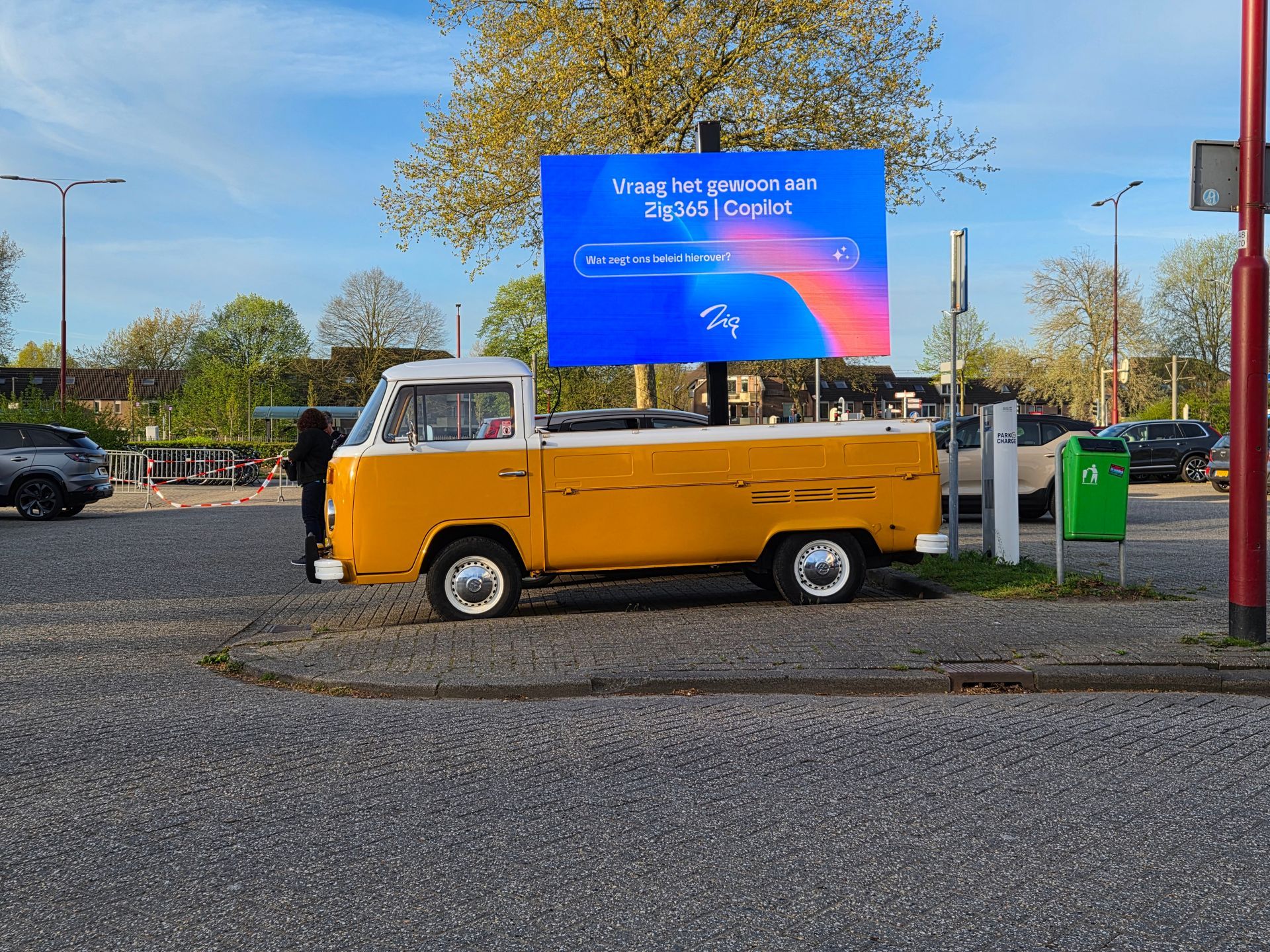 Een geel vintage busje staat geparkeerd naast een reclamebord langs de weg op een zonnige dag.