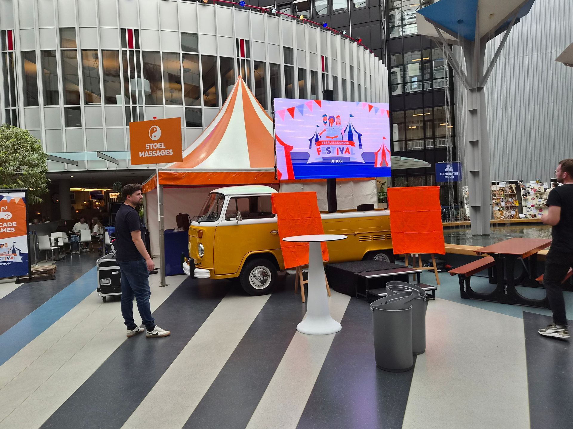 Buitenplein met een oranje volkswagen ledscherm busje, mensen zitten aan tafels in een binnenruimte.