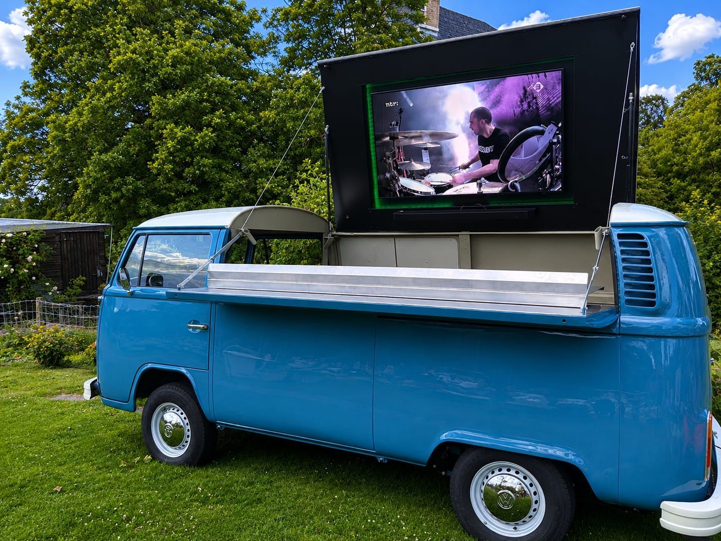 Blauwe foodtruck met een scherm op het dak waarop een optreden te zien is in een zonnig, grasrijk park.