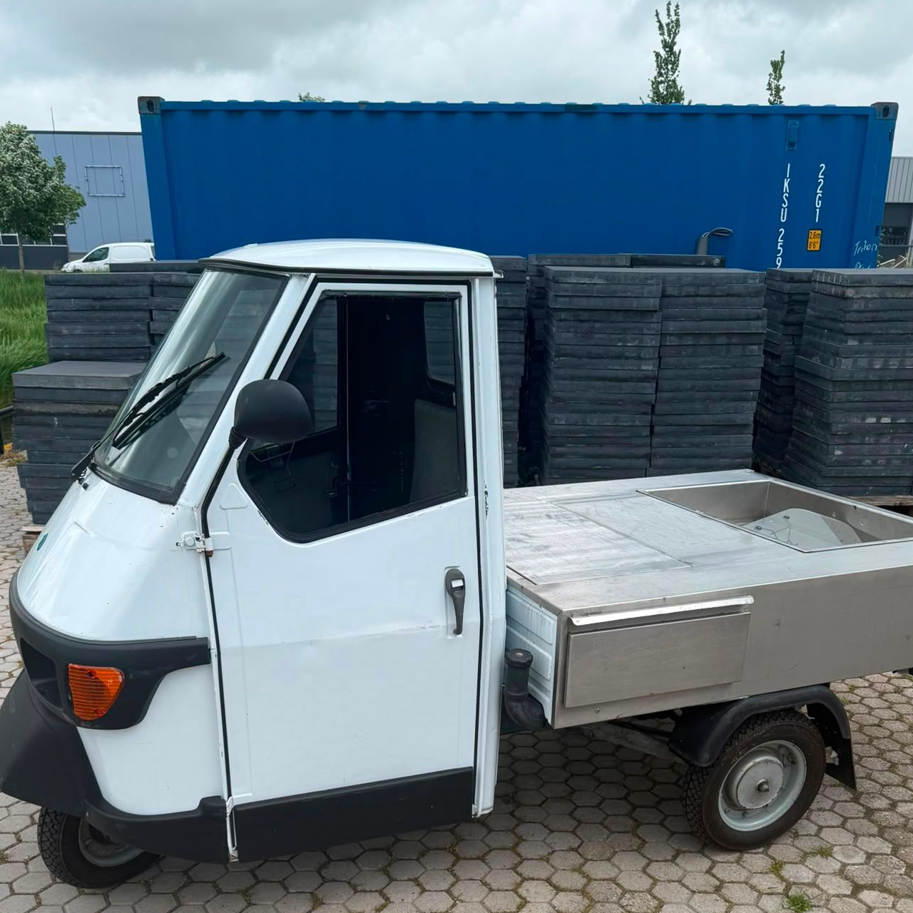 Een kleine witte driewieler staat geparkeerd naast gestapelde pallets en een blauwe zeecontainer buiten.