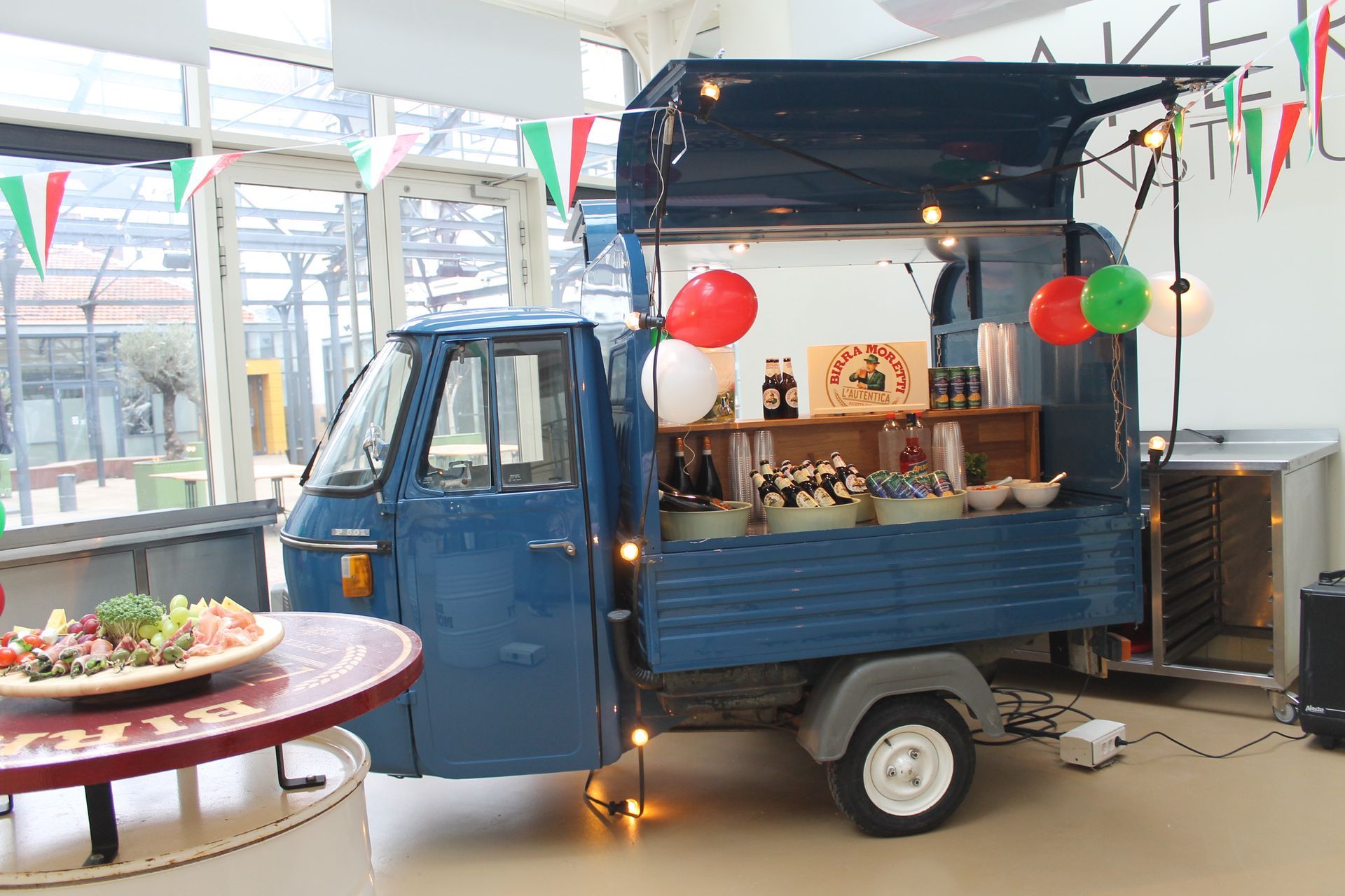 Een blauwe foodtruck op een evenement, versierd met rode en groene ballonnen, serveert eten onder een open luifel.