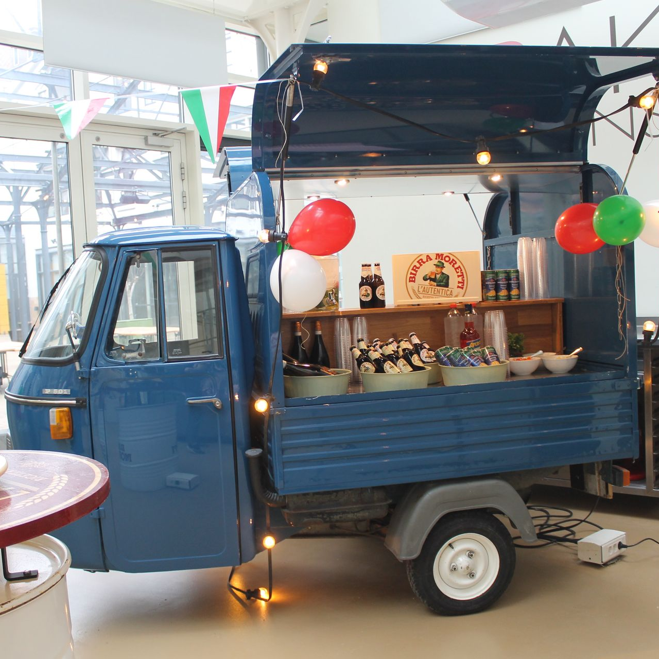Blauwe foodtruckkraam versierd met lichtslingers en ballonnen, met producten uitgestald op schappen binnen.