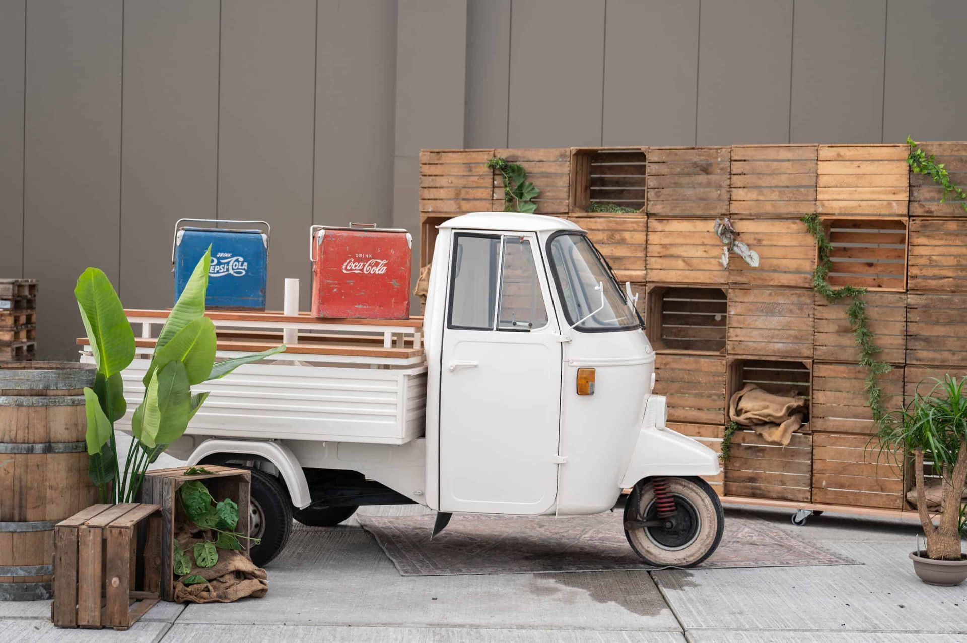 Een witte tuktuk met laadbak staat geparkeerd naast groene planten, met rode en blauwe decorstukken.