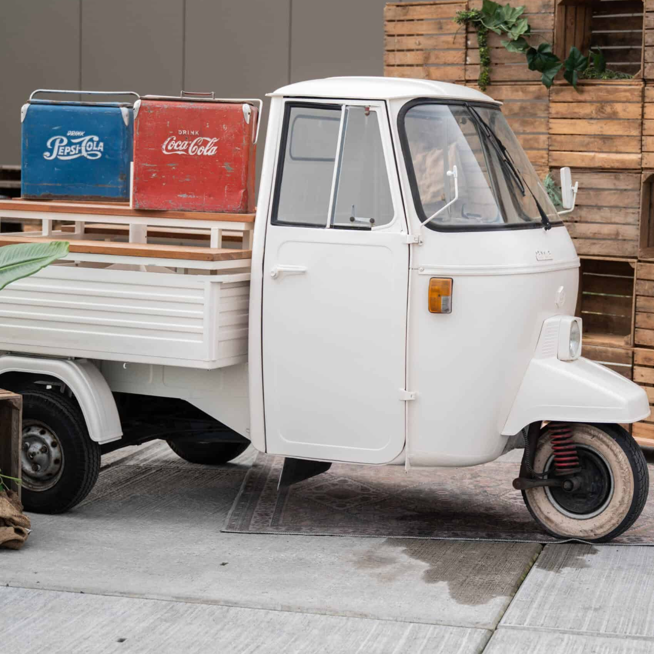 Een kleine witte tuktuk staat geparkeerd bij een bakstenen muur en vervoert rode en blauwe drankkratten.