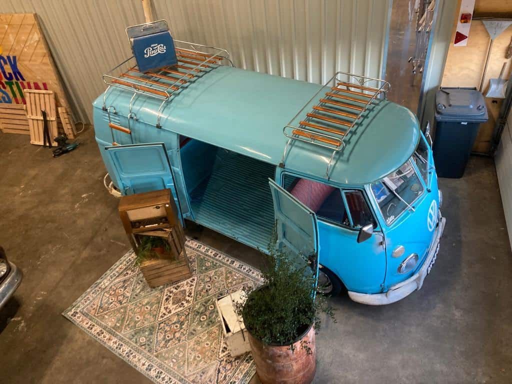 Een blauwe retro volkswagen bus staat tentoongesteld in een magazijn, met een imperiaal en planten eromheen.