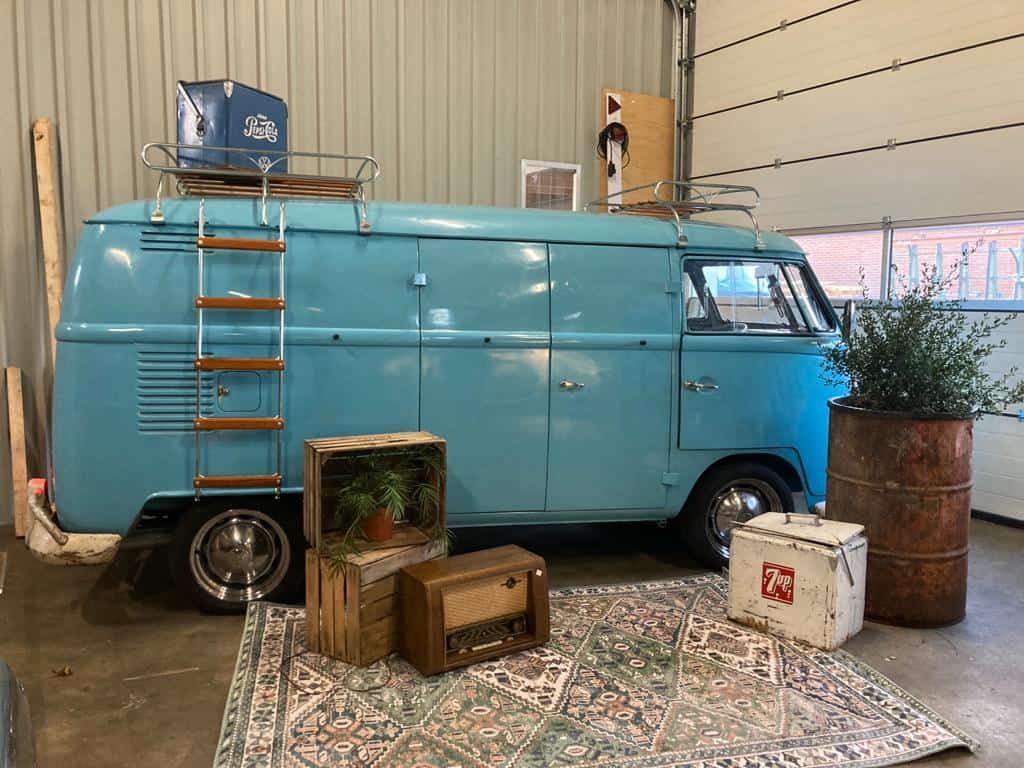 Turquoise vintage bestelwagen in een binnenvitrine met gestapelde bagage en een plantenbak van een vat.