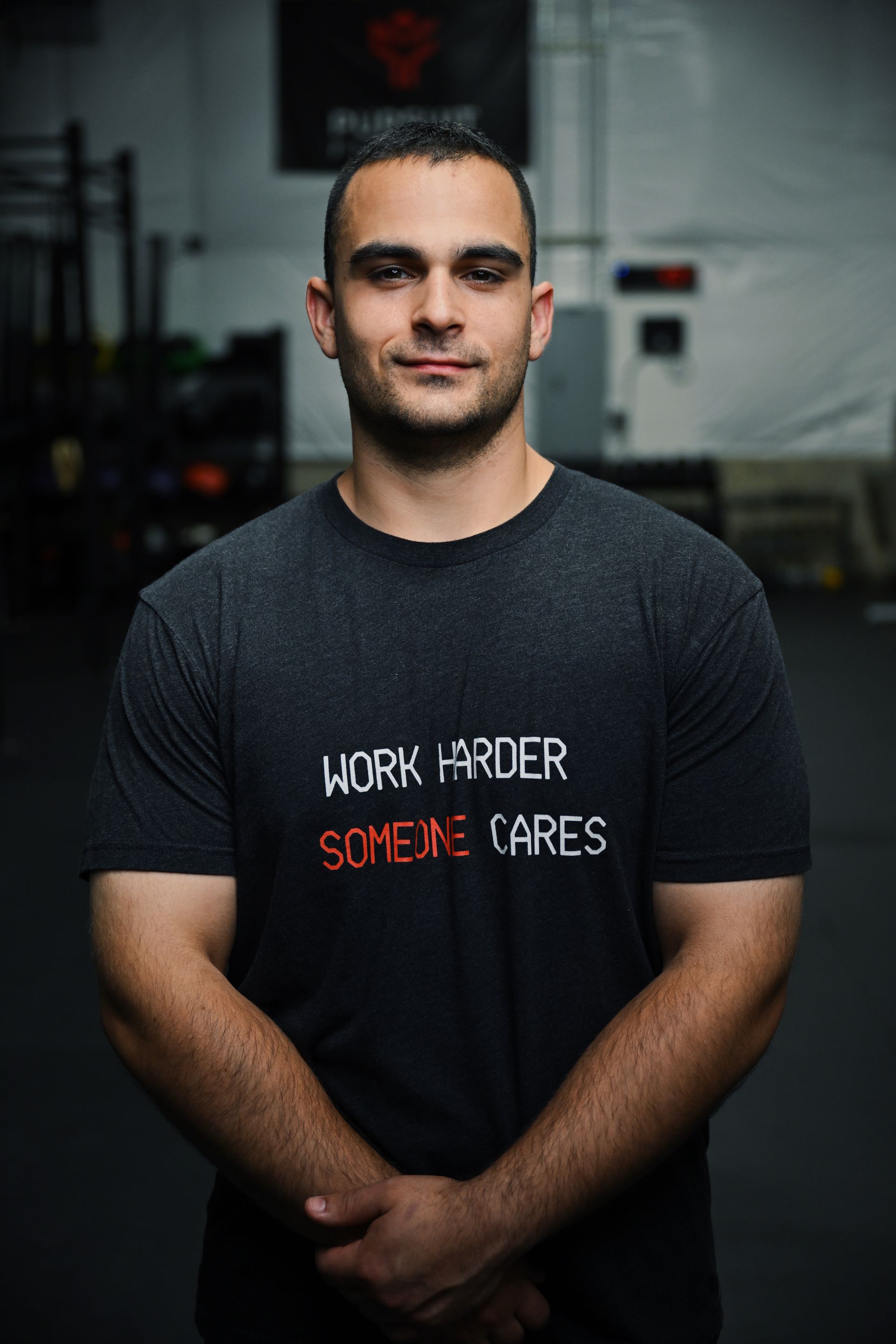 Man standing indoors in a black T-shirt with “Work Order Science Games” text, hands clasped.