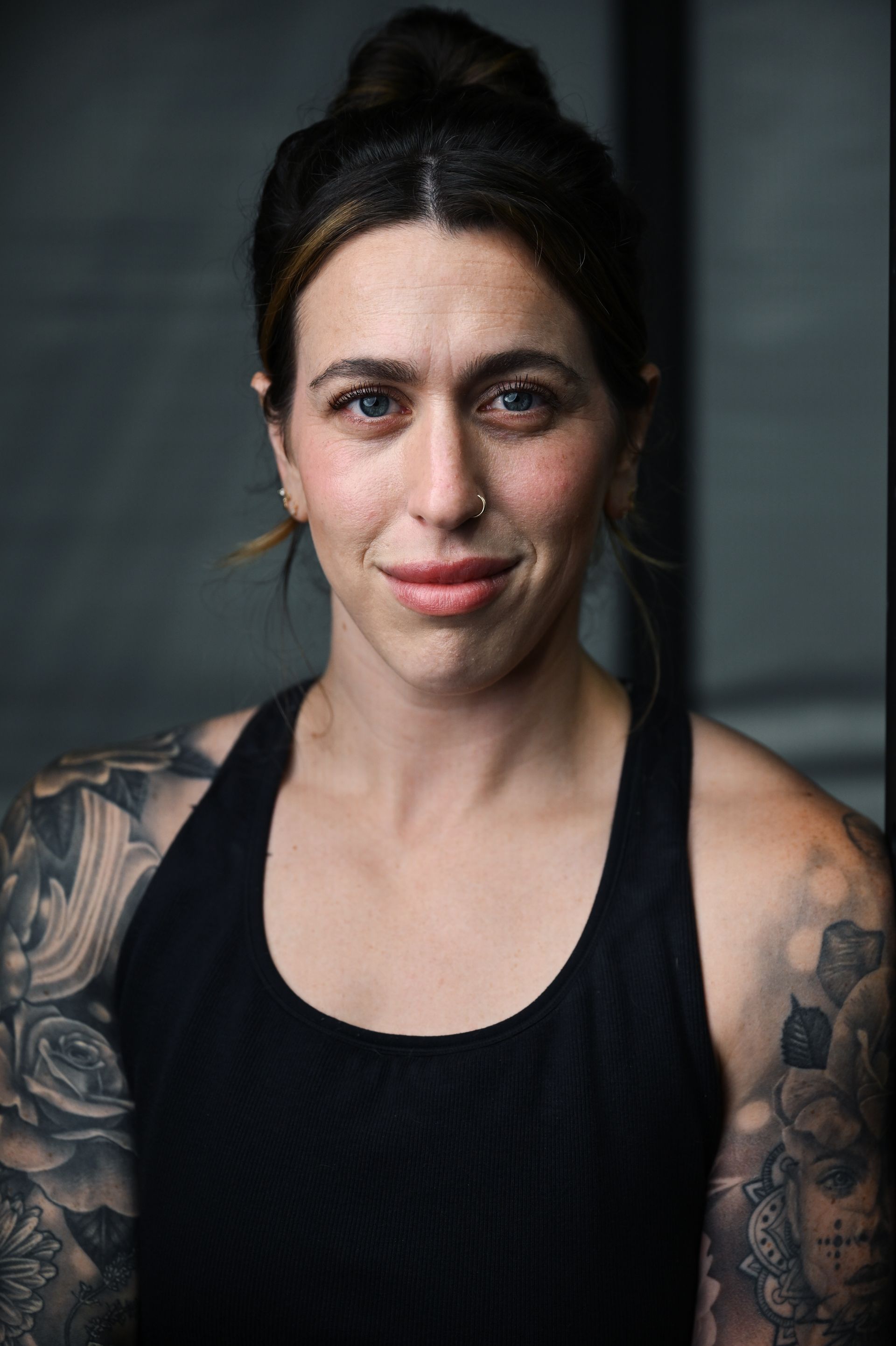 Portrait of a person with dark hair in a black tank top, showing tattooed arms, against a dark background