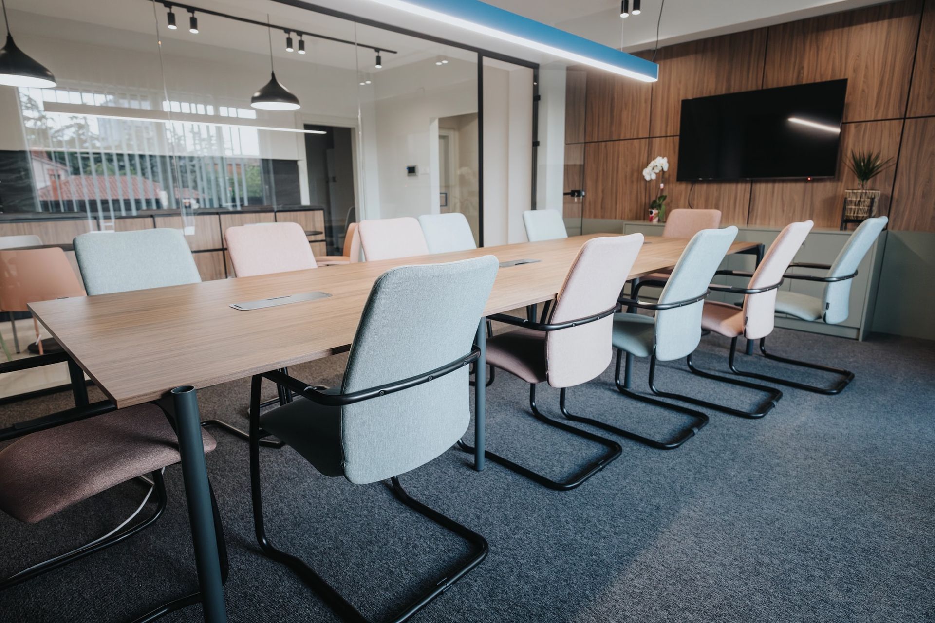 A modern conference room with a long wooden table and colorful chairs. A large TV hangs on a wood-paneled wall.