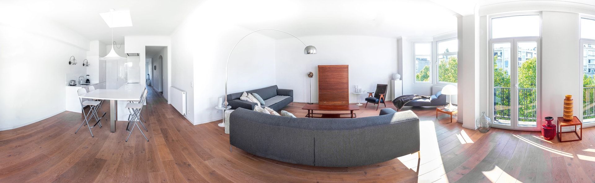 Panoramic view of a modern living room with gray sofas, wooden floors, and large windows overlooking greenery.