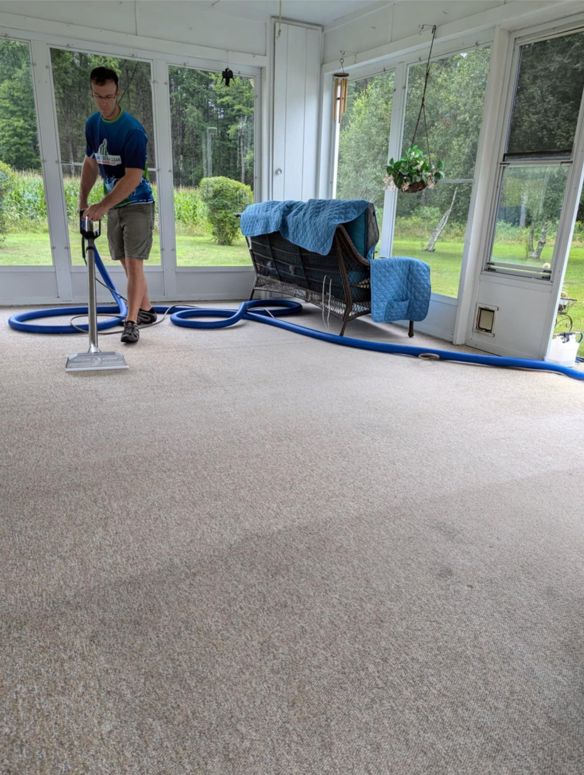 Man using a floor cleaner on beige carpet in a bright sunroom with large windows.