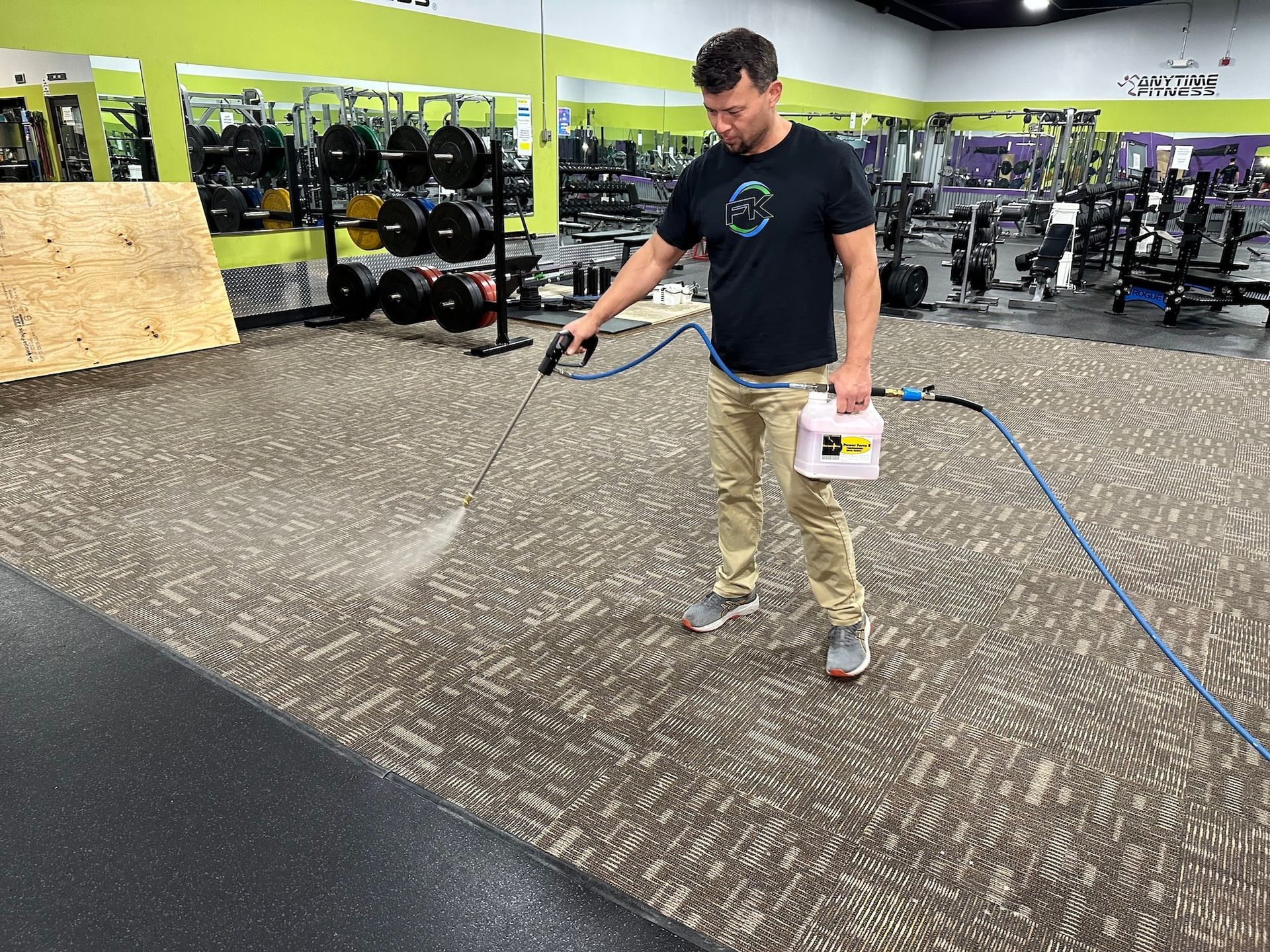 Man spraying disinfectant on the carpet in a gym. He holds a sprayer and wears a dark shirt and khaki pants.