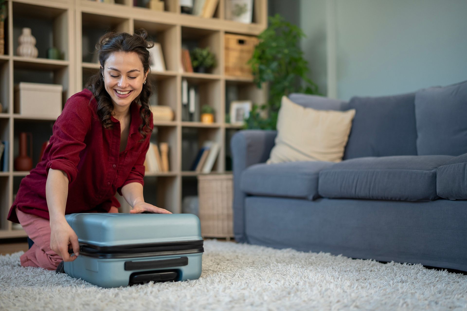 Woman kneeling on a rug with a suitcase in a cozy living room, smiling as she packs or unpacks it.