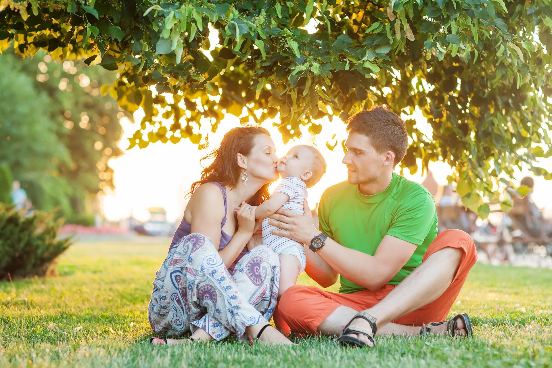 A mom, dad and child are sitting in a park. The mother is kissing her baby.