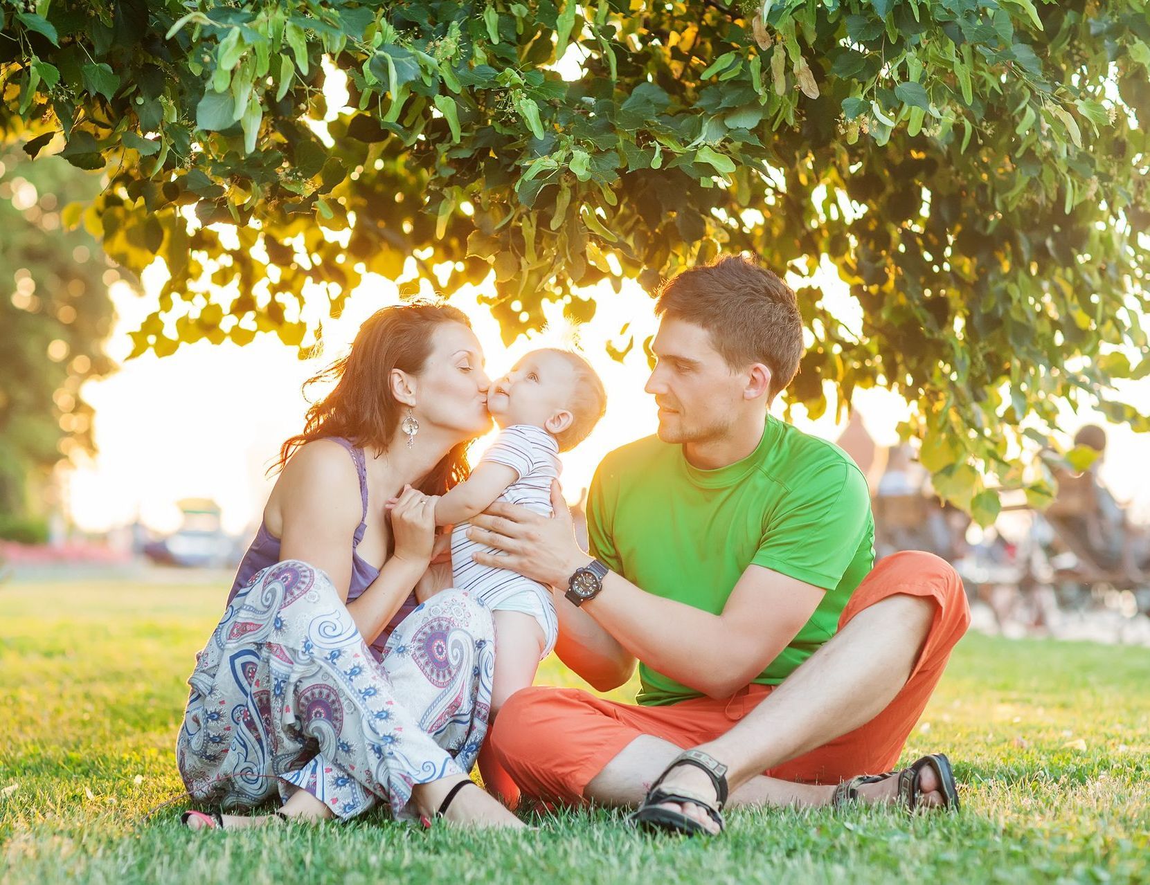 Mother and father with a baby, sitting in the park. The mother is kissing her young baby.