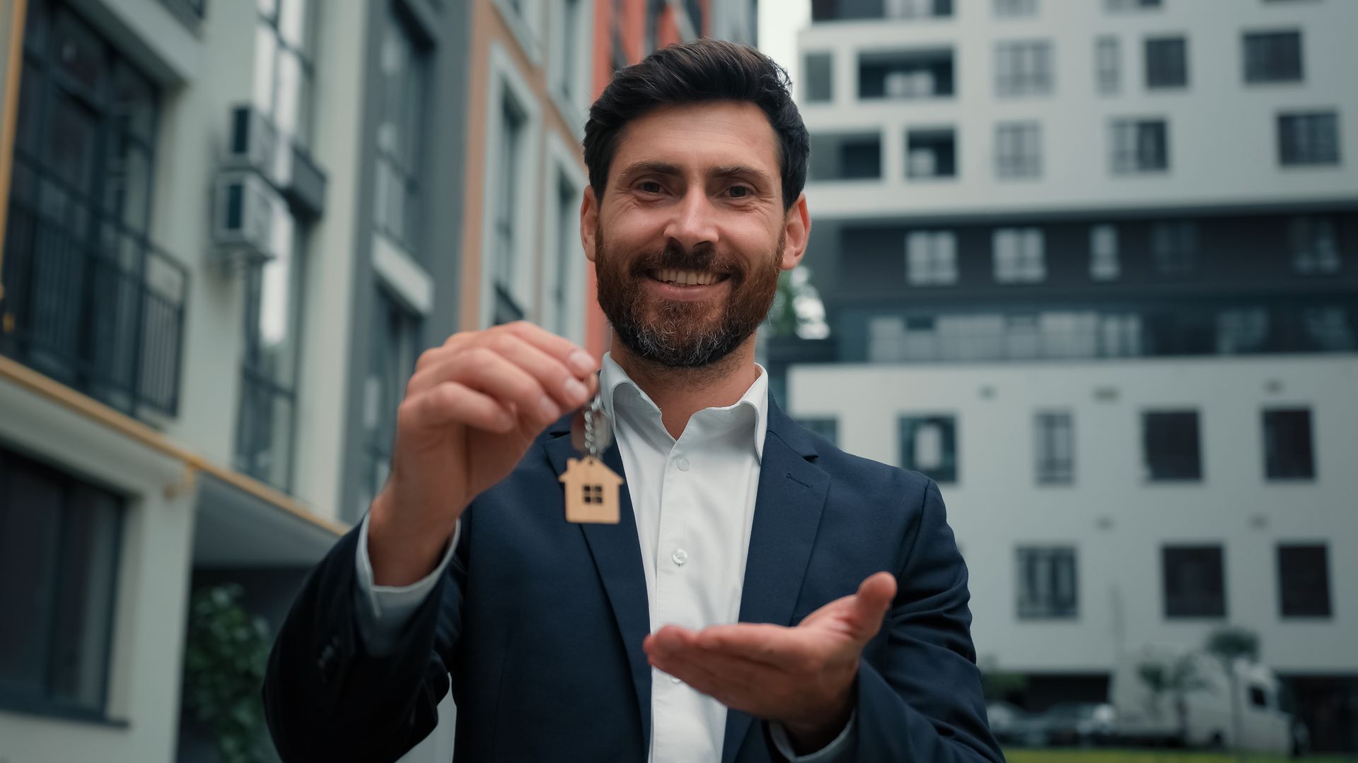 Smiling man holding up house keys in front of an apartment building