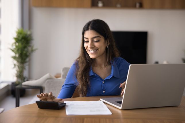 Woman smiling at a laptop on a wooden table with papers and a bowl in a bright living room