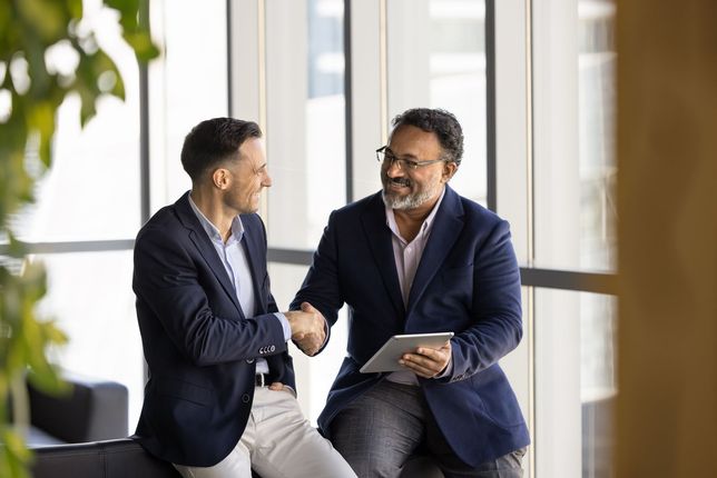 Two colleagues in blazers shaking hands and smiling in a bright office lobby