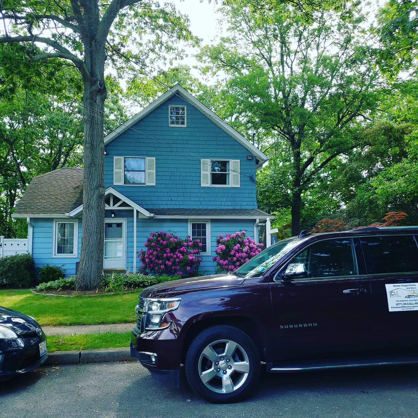 Blue house with white trim and pink flowers, partially blocked by parked SUVs on a tree-lined street