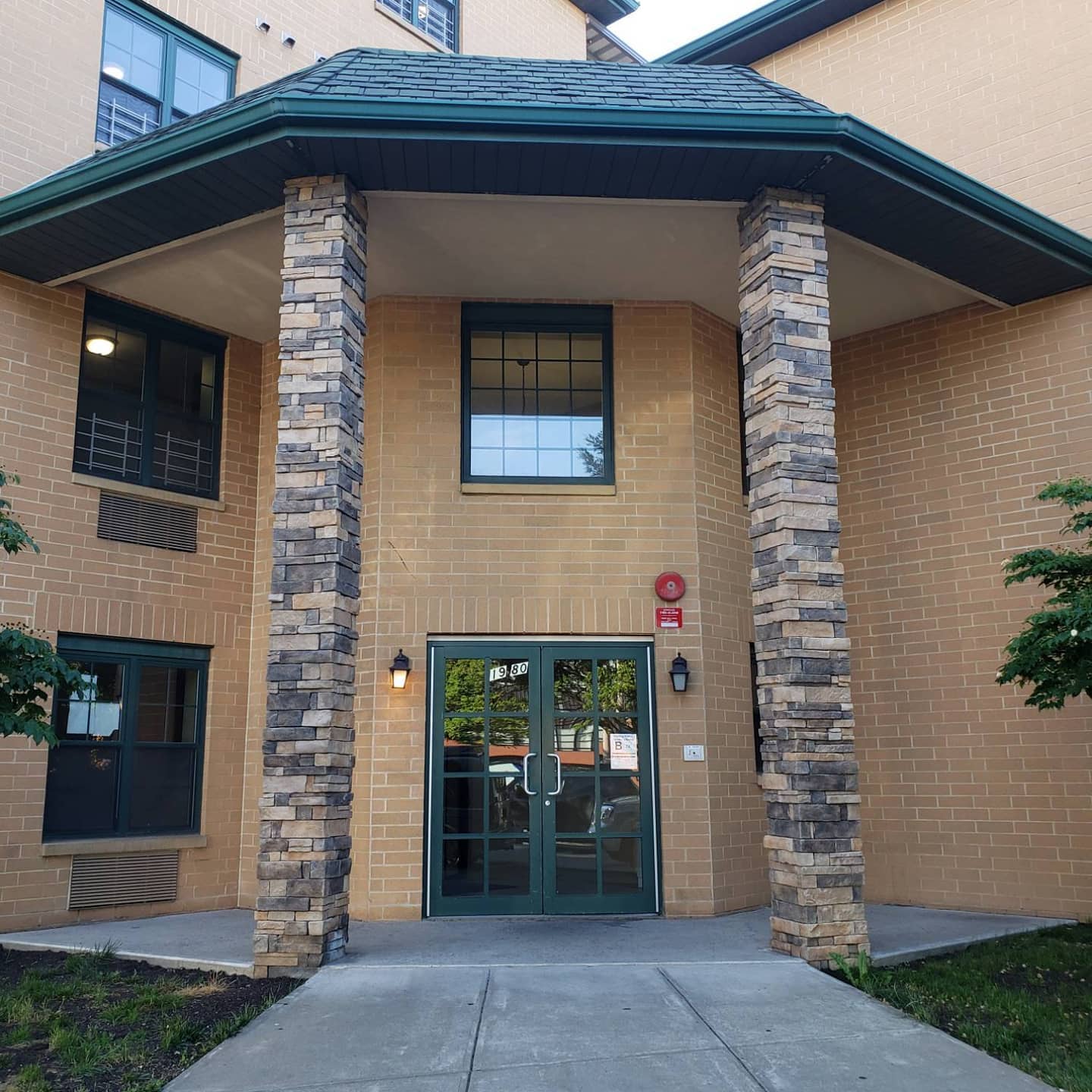Building entrance with stone columns, tan brick walls, and glass double doors under a dark green roof.