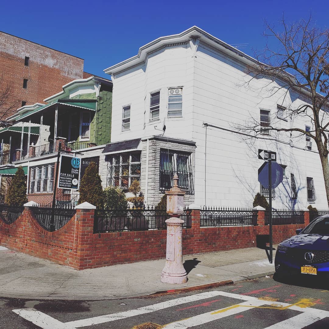 White corner house with brick fence on a sunny street, parked car at right and traffic sign nearby