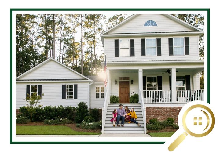 White farmhouse and garage with porch, trees behind, and family sitting on front steps
